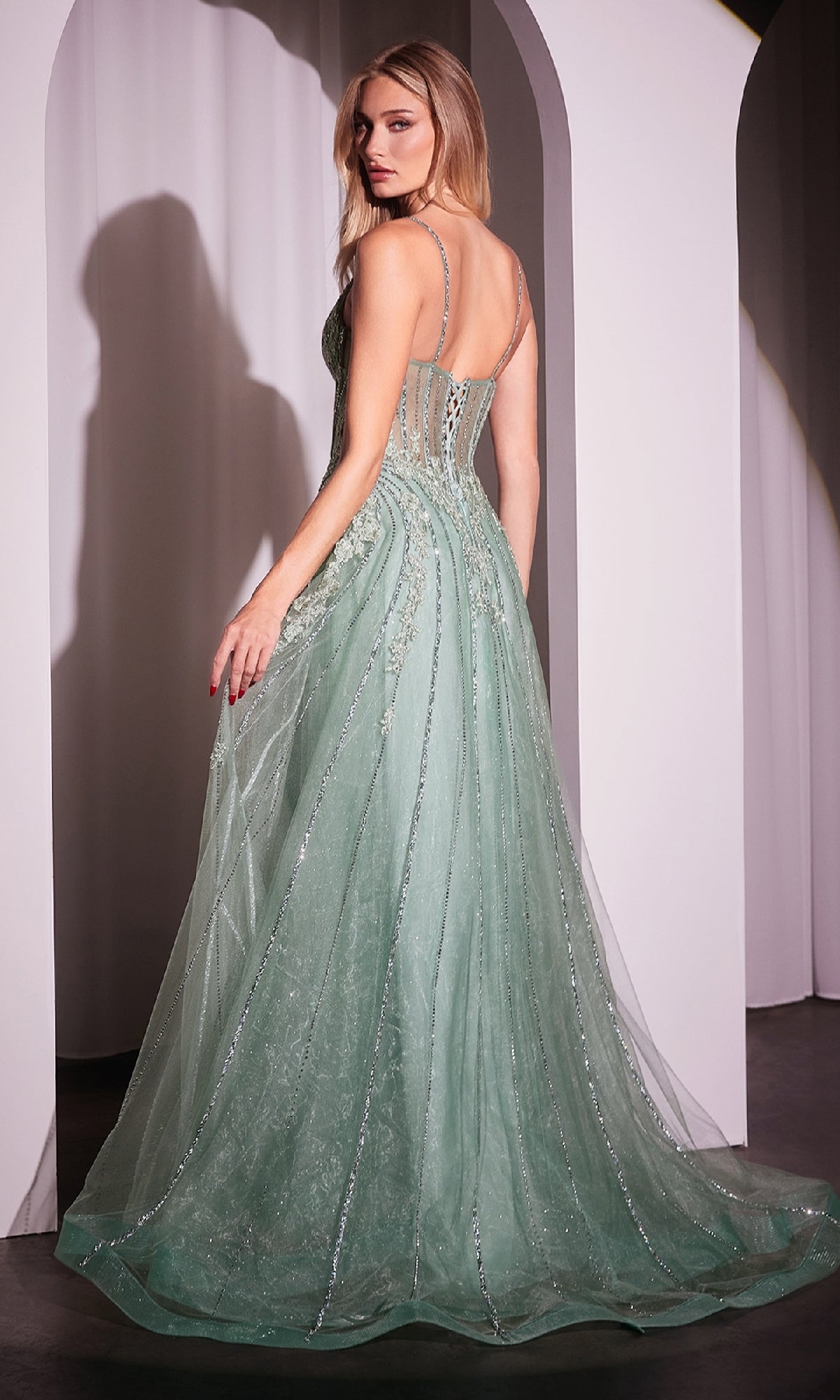 A woman stands with her back turned, looking over her shoulder in the Ladivine CD786 Crystal-Beaded Long Prom Dress—a light green, floor-length gown featuring sheer panels and intricate embellishments.